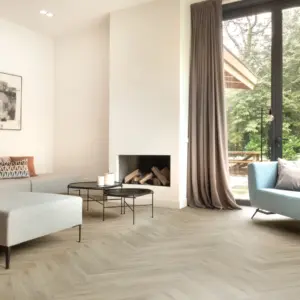 Sfeervolle woonkamer met een lichte greige eiken Hebeta HB visgraat pvc-vloer. De ruimte beschikt over een strakke witte schouw met een ingebouwde houthaard, twee ronde zwarte salontafels, een grijze zithoek en een lichtblauwe designbank bij een grote glazen pui met lange taupe gordijnen.