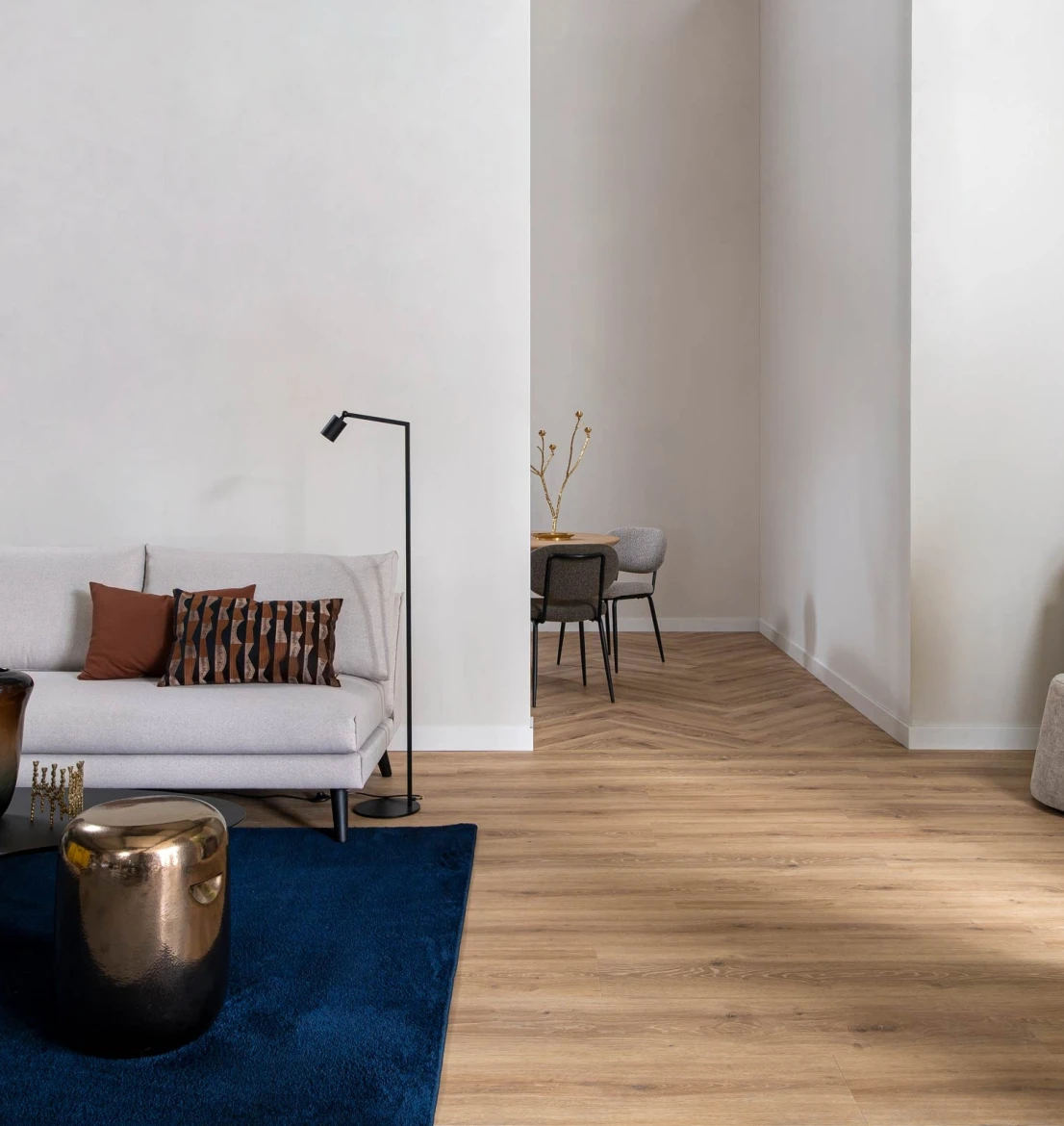 Modern interieur met een Hebeta HB Chablis XL pvc-vloer. Op de voorgrond liggen rechte houtlook planken bij een grijze bank met blauw vloerkleed. In de achterliggende eetkamer loopt de vloer drempelloos over in een visgraat pvc-patroon.