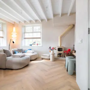 Sfeervol interieur met een lichte naturel eiken Hebeta HB visgraat pvc-vloer. De ruimte is ingericht met twee halfronde beige stoffen banken, een wit balkenplafond, een kleine beige houtkachel en zachte zandkleurige wanden met veel daglicht.