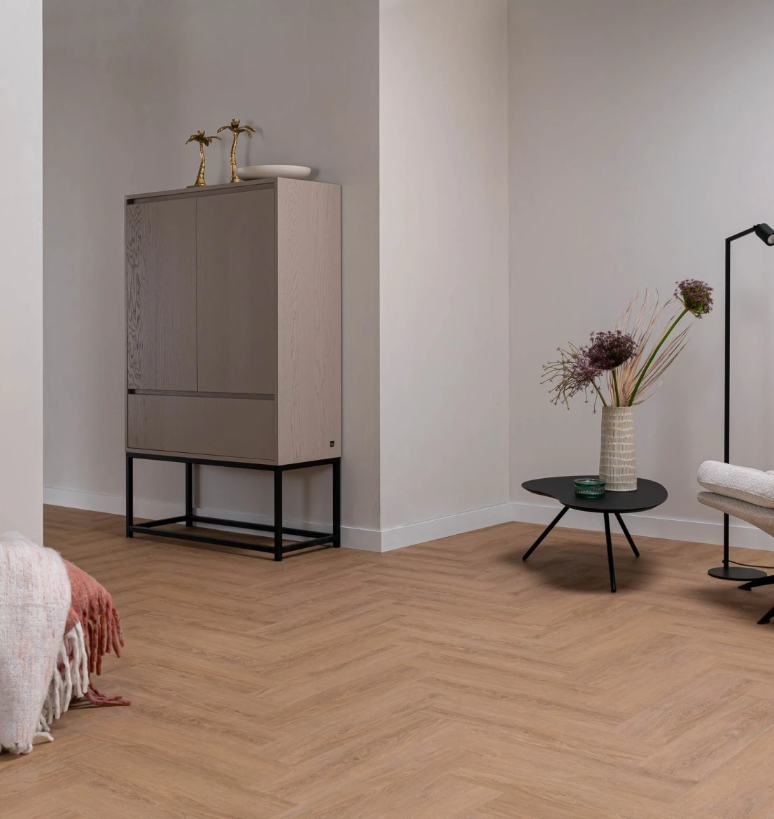 Warm eiken visgraatvloer in een modern, licht interieur met een minimalistische grijze designkast op een zwart onderstel en een zwarte salontafel met droogbloemen.