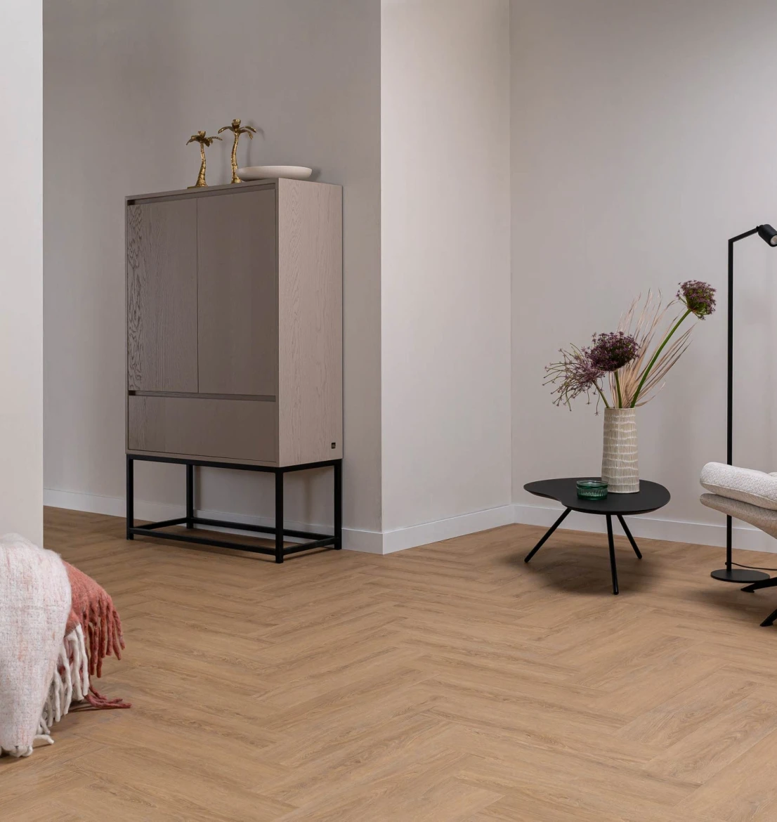 Sfeervol, modern interieur met een warm eiken visgraat PVC vloer uit de HB Charente Visgraat XL collectie. Links staat een hoge lichtgrijze designkast op een zwart metalen frame met gouden decoratie. Rechts staat een organische zwarte salontafel met een beige vaas en droogbloemen naast een witte loungestoel.