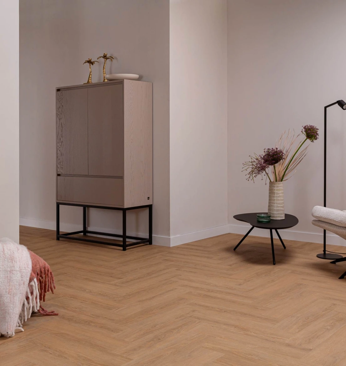 Eigentijdse woonkamer met een natuurgetrouwe HB Charente Visgraat XL PVC vloer in warm eiken houtlook. In de lichte ruimte staat een minimalistische grijze opbergkast op een zwart stalen frame en een ronde zwarte salontafel met een vaas sierlijke droogbloemen.