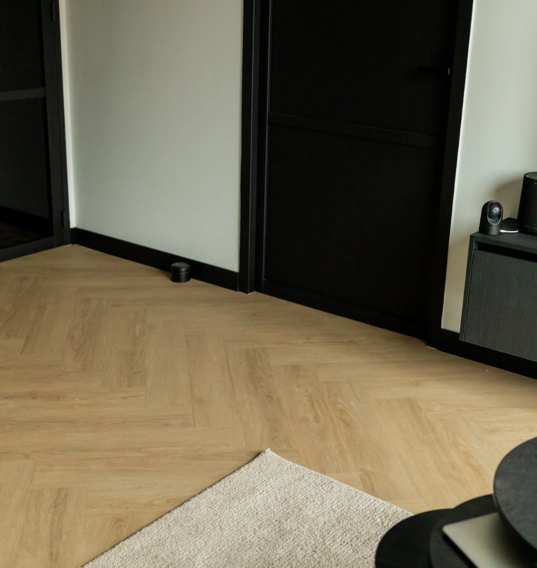 Modern interieur met een lichte eiken PVC visgraatvloer uit de HB Charente collectie. De vloer contrasteert met strakke witte muren, een matzwarte deur met bijpassend zwart kozijn en zwarte plinten. Op de voorgrond is de hoek van een beige vloerkleed en een zwarte salontafel zichtbaar, en rechts hangt een donker zwevend tv-meubel.