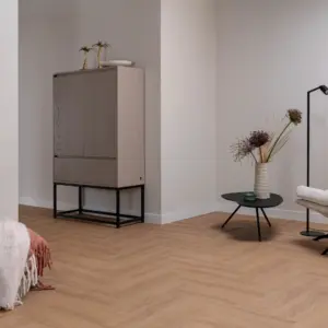 Sfeervol interieur met de Hebeta HB visgraat pvc-vloer in warm eiken. Tegen een strakke greige muur staat een hoge, lichtgrijze houten kast met zwart onderstel. Rechts staat een ronde zwarte bijzettafel met een gestructureerde vaas met droogbloemen en een zwarte vloerlamp.
