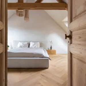 Blik door een open houten deur naar een lichte slaapkamer met een Hebeta HB visgraat pvc-vloer in naturel eiken. De kamer heeft rustieke houten dakbalken, een beige gestoffeerd tweepersoonsbed, een houten nachtkastje en zachte, kalkwitte wanden onder een schuin dak.