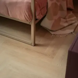 Sfeervolle close-up van een lichte eiken PVC visgraatvloer in een slaapkamer. Links staat het beige metalen frame van een bed met oudroze beddengoed. Rechts hangt een wollen, zachtroze met beige plaid met franjes deels over de vloer vanaf het bed. Rechtsvoor is een onscherpe hoek van een donkerpaars meubel zichtbaar.