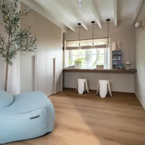 Sfeervolle kamer met een warm naturel eiken Hebeta HB Château XL Plank pvc-vloer. De ruimte heeft een wit houten balkenplafond en een ingebouwde beige kastenwand. Onder het raam bevindt zich een houten bureau-werkblad met twee witte krukjes en drie hanglampen, met op de voorgrond een lichtgrijze of lichtblauwe zitzak en een kunstolijfboom.
