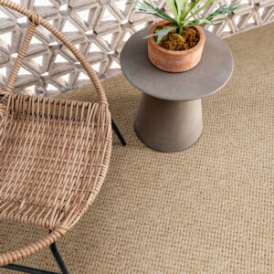 Sfeervol interieur of terras met een goudbruin, fijn geweven sisal-look tapijt als vloer, gecombineerd met een rotan stoel en een betonnen bijzettafel met plant.