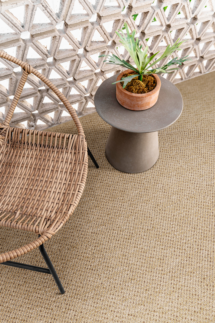 Sfeervol interieur of terras met een goudbruin, fijn geweven sisal-look tapijt als vloer, gecombineerd met een rotan stoel en een betonnen bijzettafel met plant.