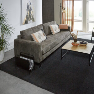 Modern woonkamer interieur met een groot, antracietzwart hoogpolig vloerkleed op een grijze betonvloer, ingericht met een stoffen hoekbank, houten salontafel en kamerplant.