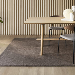 Een sfeervolle interieurfoto in een minimalistische Japandi-stijl, waarin een rechthoekig, taupekleurig gemêleerd vloerkleed centraal op de grond ligt. Het kleed heeft een dichte, comfortabele pool met een subtiele textuur van gemengde bruin-, grijs- en taupetinten.Het vloerkleed ligt op een lichte eikenhouten vloer. Een grote, bijpassende eikenhouten eettafel staat deels op het kleed, omringd door moderne eetkamerstoelen met donkere, slanke metalen poten en halfronde houten rugleuningen met een grijze stoffen zitting. Op de achtergrond bevindt zich een kamerhoge wand bekleed met verticale, houten akoestische latten (woodpanels) in een warme eikenkleur, wat zorgt voor een prachtig lijnenspel. Links in de hoek staat een grote, groene kamerplant (Strelitzia) in een grijze pot. Op het rechtergedeelte van de tafel staan minimalistische, keramische vazen en decoraties in wit- en zandkleuren. De ruimte baadt in zacht, natuurlijk daglicht, wat de rustige en warme sfeer van het tapijt accentueert.