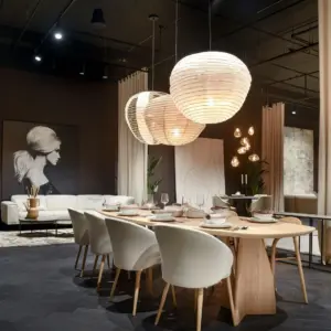 Luxe, modern interieur met een donkergrijze tot zwarte eiken PVC visgraatvloer. Centraal staat een grote, ovale houten eettafel die sfeervol is gedekt met servies, omringd door zes beige design eetkamerstoelen met houten pootjes. Boven de tafel hangen twee grote, bolvormige papieren design hanglampen. Op de achtergrond bevindt zich een zithoek met een lichte bank, een rond salontafeltje en een groot zwart-wit portret van een vrouw aan een donkere wand.