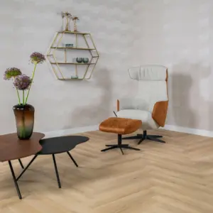 Sfeervolle hoek in een kamer met de Hebeta HB visgraat pvc-vloer in een lichte naturel eikenkleur. De ruimte is ingericht met een beige met cognac design fauteuil en bijbehorende hocker, een set organisch gevormde salontafels met een vaas paarse Alliums, en een zeshoekig goudkleurig wandschap tegen een beige behangwand.
