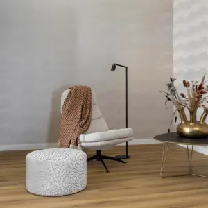 Sfeervolle hotel-chique zithoek met de Hebeta HB Progress XL Plank pvc-vloer in een matte grijs-beige natuureiken kleur. De ruimte is ingericht met een lichte design fauteuil met bruin plaid, een ronde poef met kiezelpatroon en een goudkleurige tulpenvaas op de salontafel tegen een wand met beige geometrisch behang.